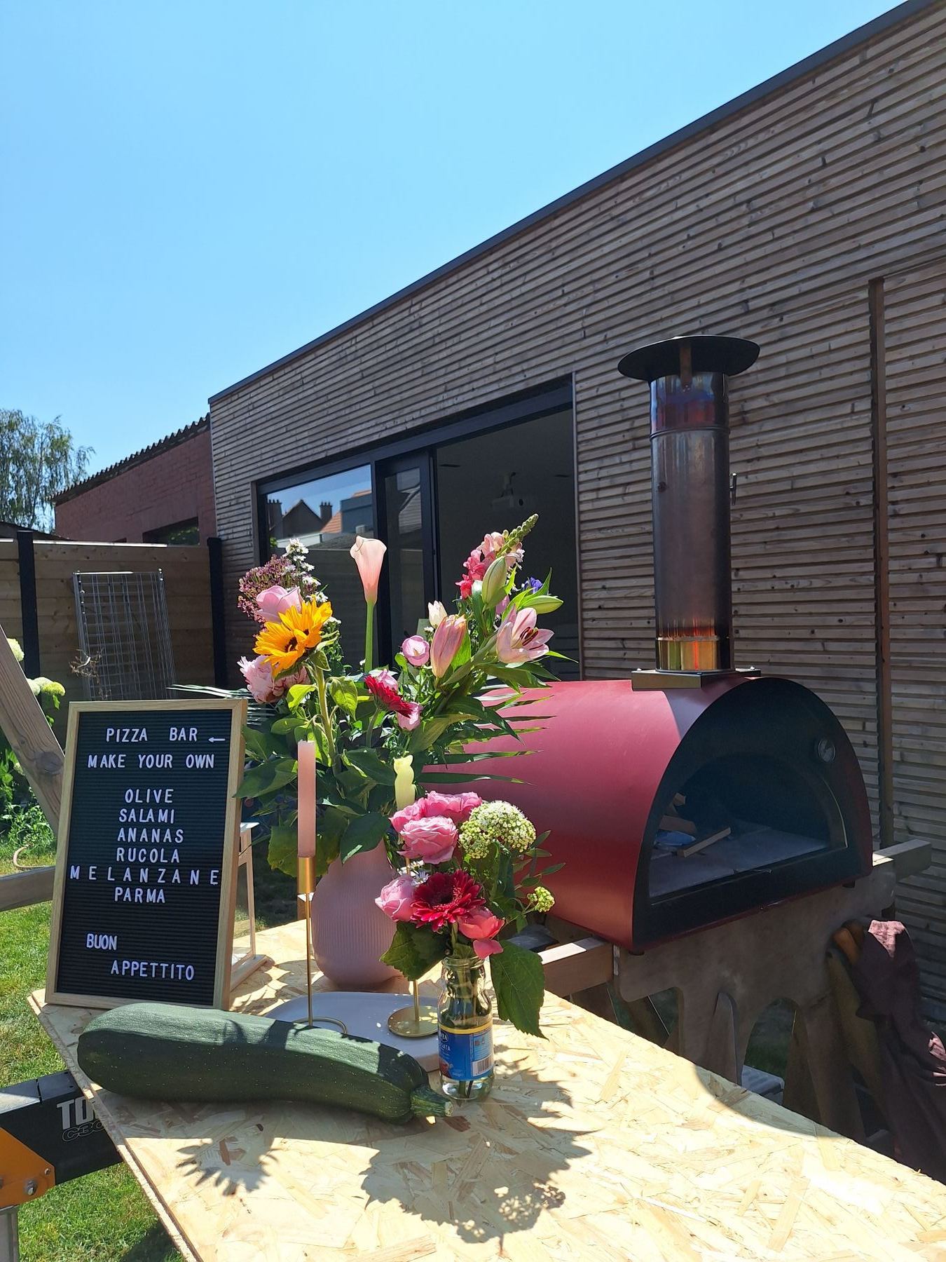 Rode houtgestookte Pizzaiolo pizzaoven met roken schoorsteen en een letterbord met het menu, opgesteld in een tuin